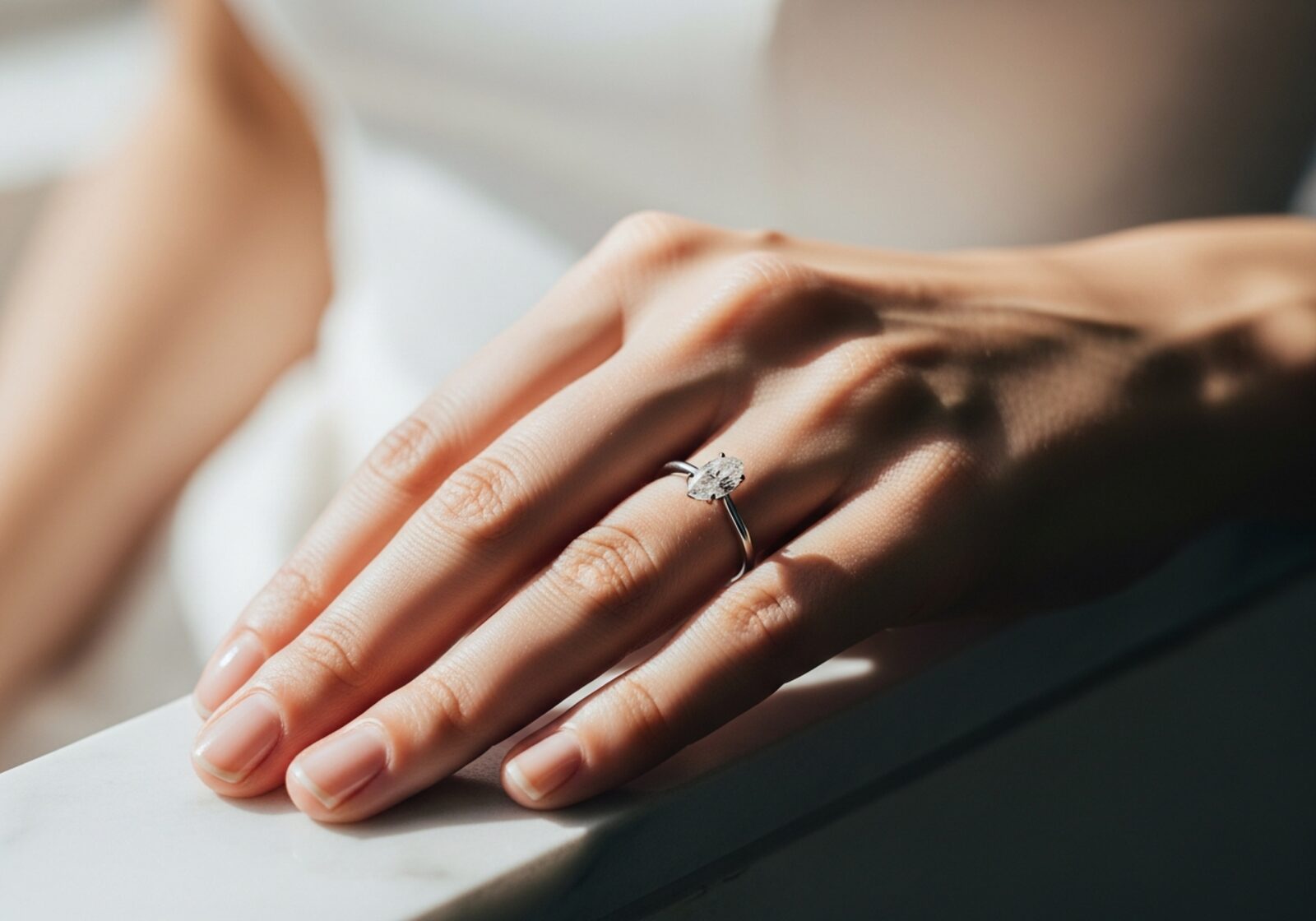 Bride's left hand resting on cream linen wearing an elongated oval diamond solitaire on a thin platinum band